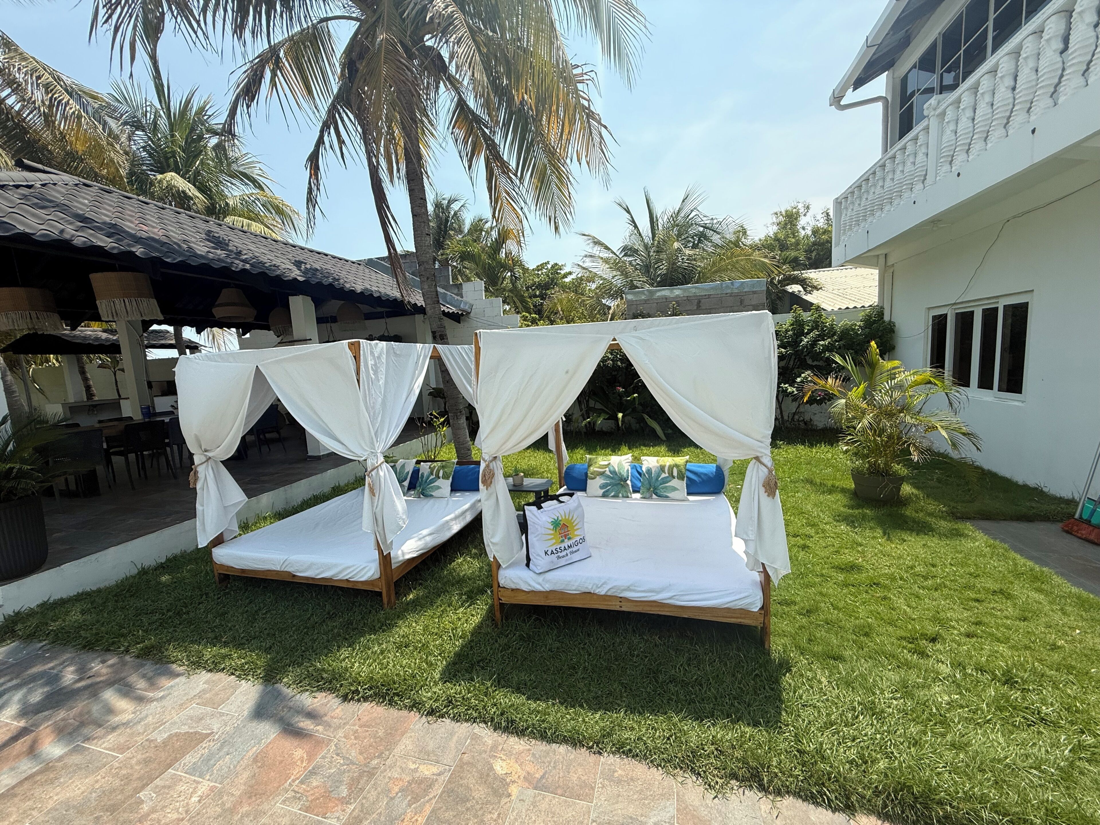 Two bed-and-breakfast-style beds with white canopies, a patio area with chairs, and a building with white walls and a balcony.