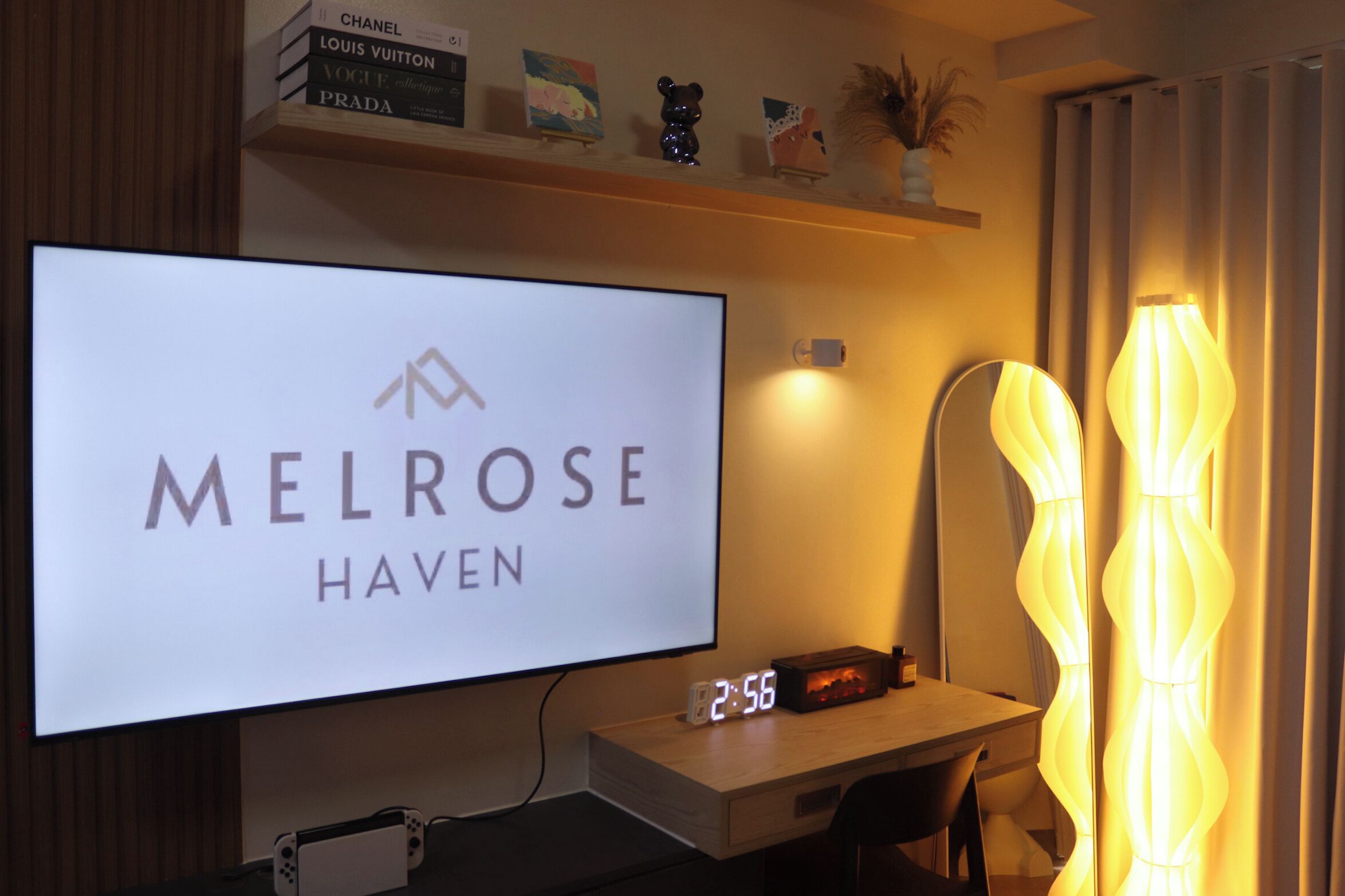 Una habitación de hotel con un televisor de pantalla plana grande mostrando 'MELROSE HAVEN' en la pared, una mesita de noche con un reloj y una lámpara decorativa.