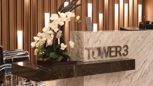 Un comptoir d’accueil avec un plateau en marbre et le nom « TOWER3 » inscrit en lettres métalliques. On peut voir un vase rempli de fleurs blanches sur le comptoir, et un fauteuil roulant est visible sur la gauche.