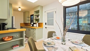 A dining area with a round table set for four, a kitchen in the background, and a window with a view of trees. - Serene Mill Valley Retreat - King Bed, Walk to Downtown, Deck, Sleeps 4. (Mill Valley)