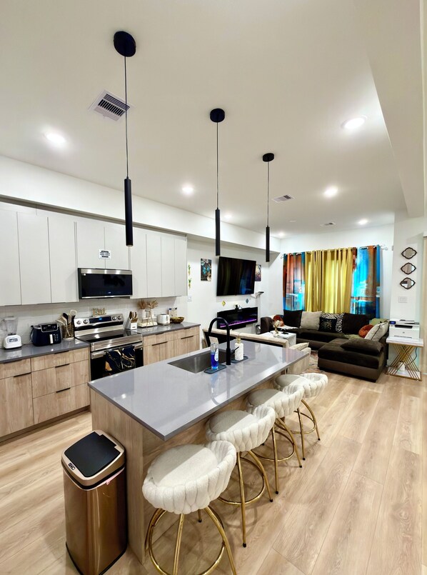 A modern kitchen with a central island, bar stools, a dining area with a sofa, and a TV mounted above the island.