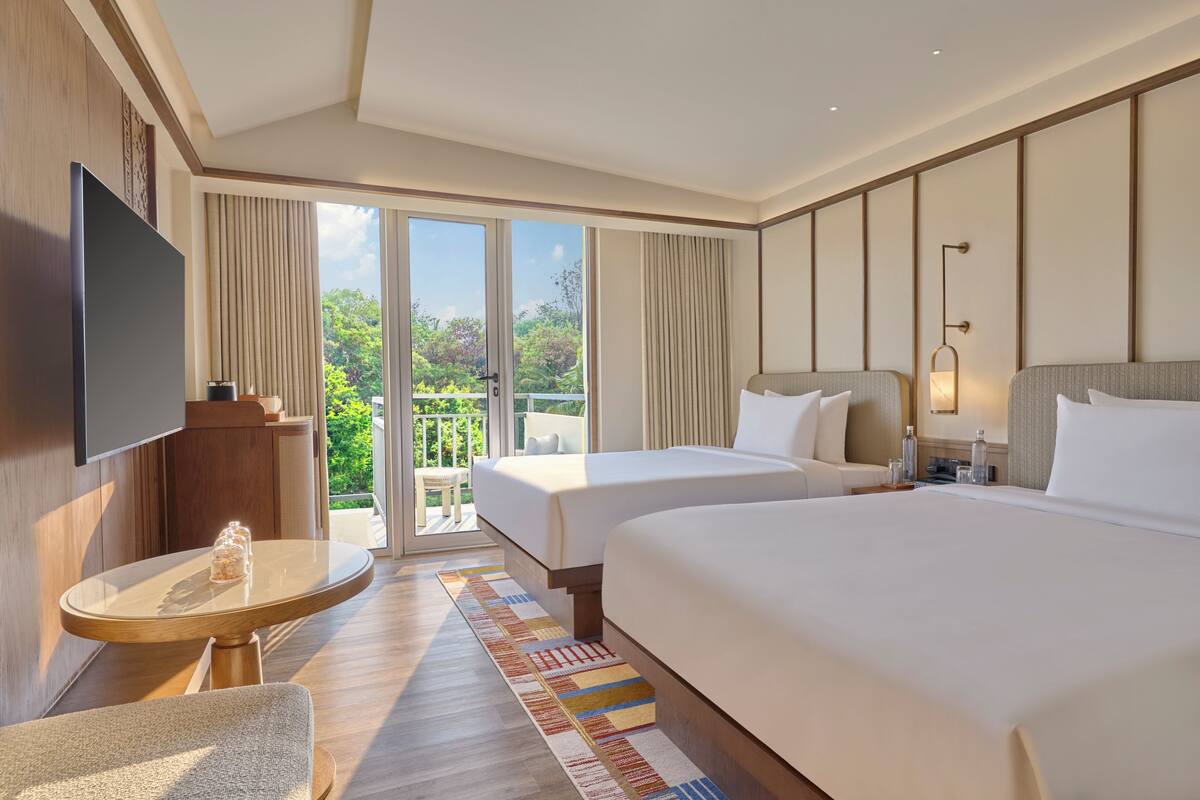 A hotel room with two beds, a TV, a balcony with a view of greenery, and a small table with a glass.