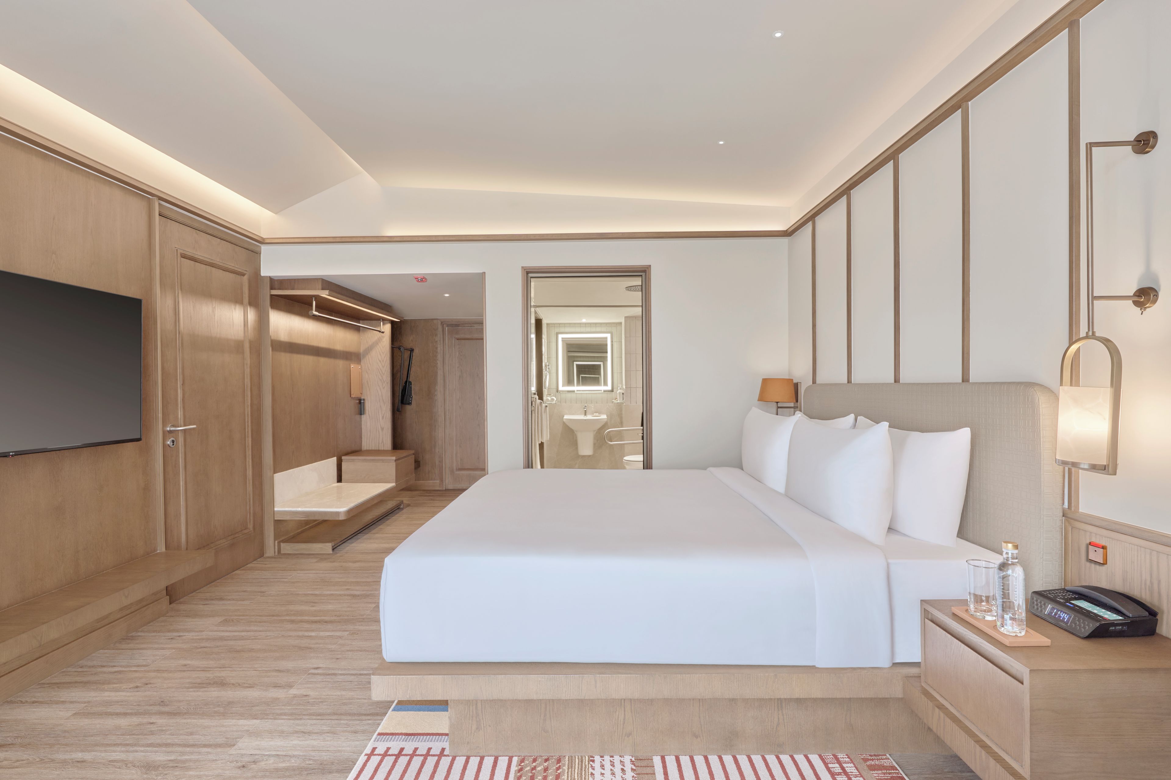 Une chambre d’hôtel avec un grand lit, une télévision, une table de chevet avec une lampe, et une salle de bain visible à travers une porte ouverte.