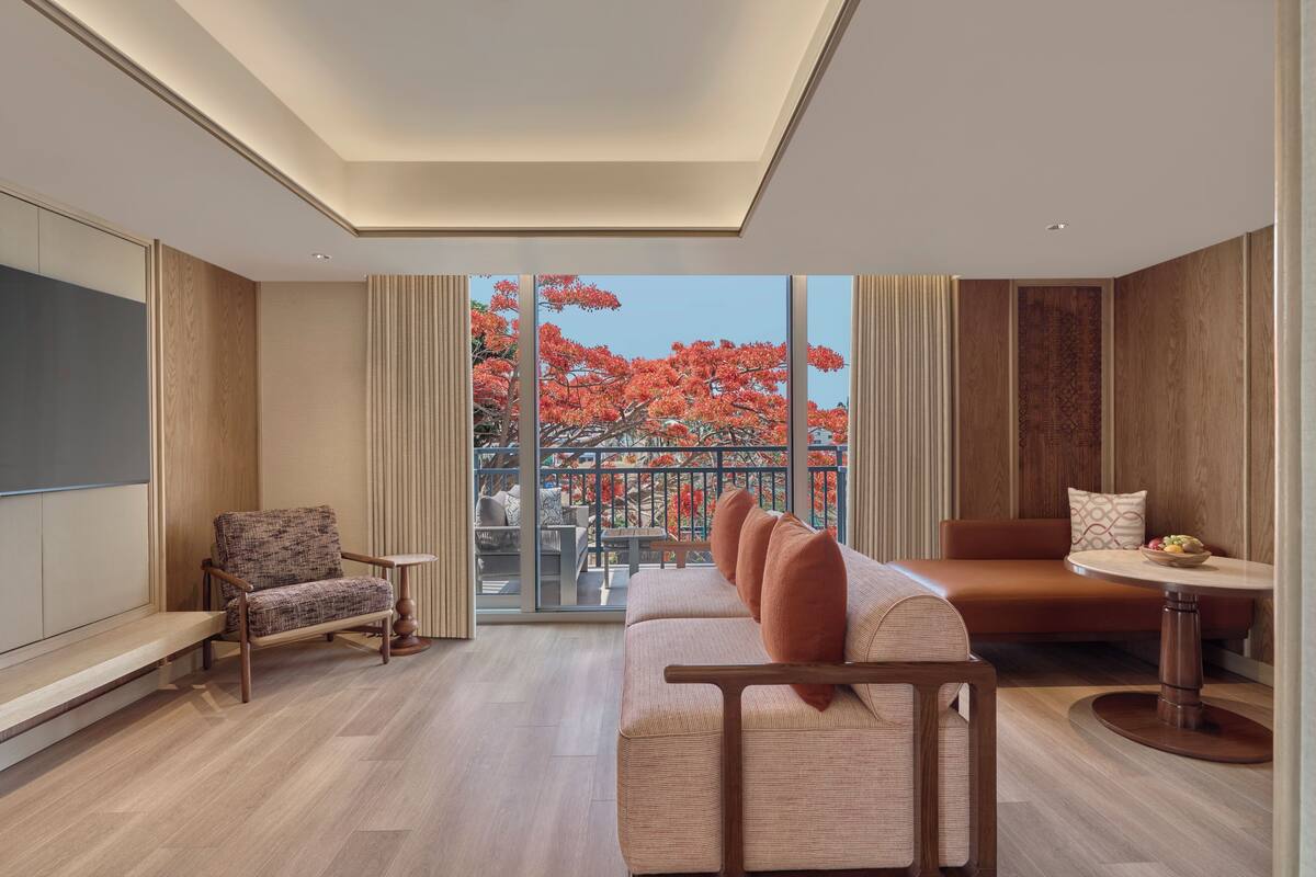A modern living room with a sofa, armchair, and round table, large windows with a view of trees, and a ceiling light.