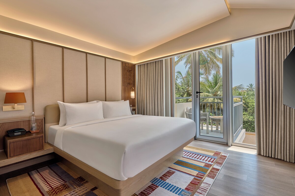 A modern hotel room with a large bed, a nightstand, a lamp, a TV, and a balcony with a view of palm trees.