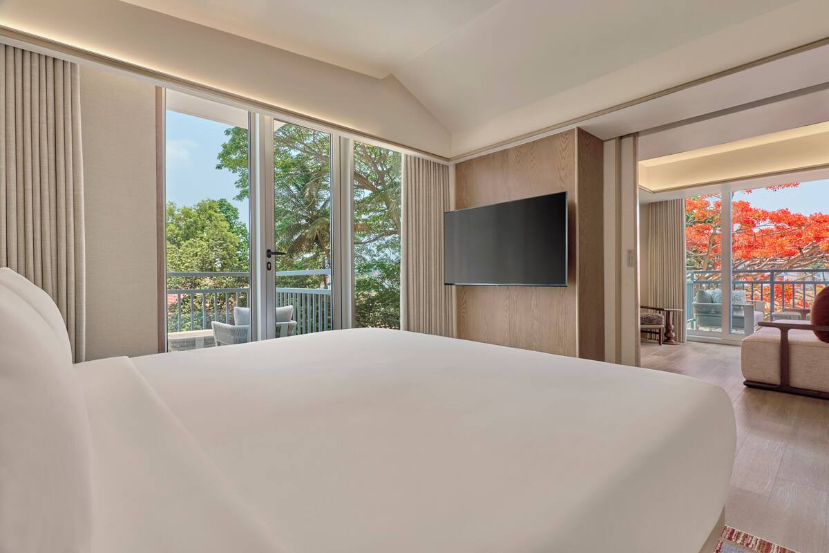 A hotel room with a large bed, a TV mounted on the wall, a balcony with outdoor seating, and a view of trees.