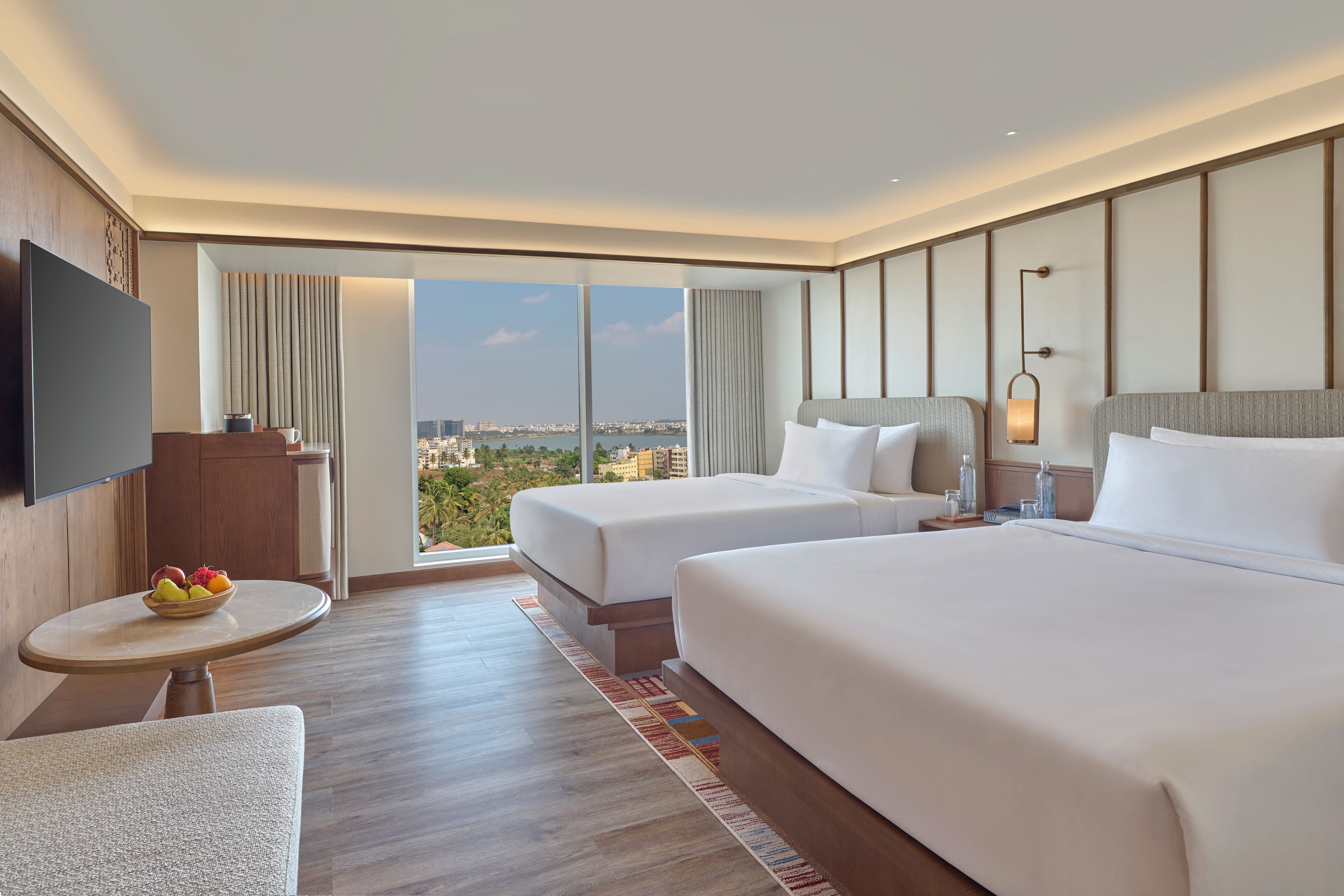 A hotel room with two beds, a TV, a small table with fruit, and a view of the city.