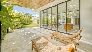 En moderne udendørs terrasse med to fletloungestole, et spiseområde med glasfacade og udsigt over en frodig have.