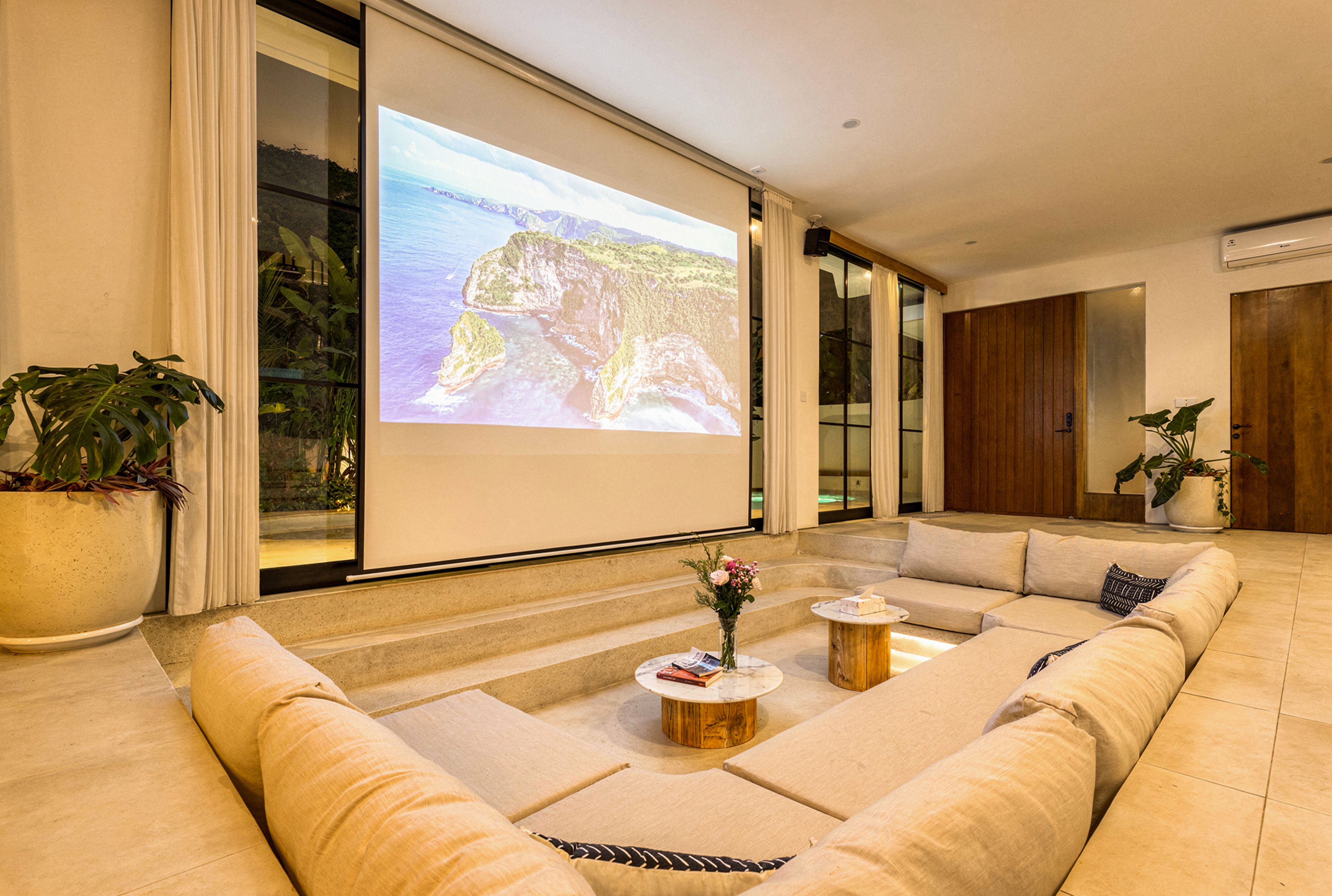 A modern living room with a large screen displaying a scenic landscape, comfortable seating, and a coffee table with a vase of flowers.