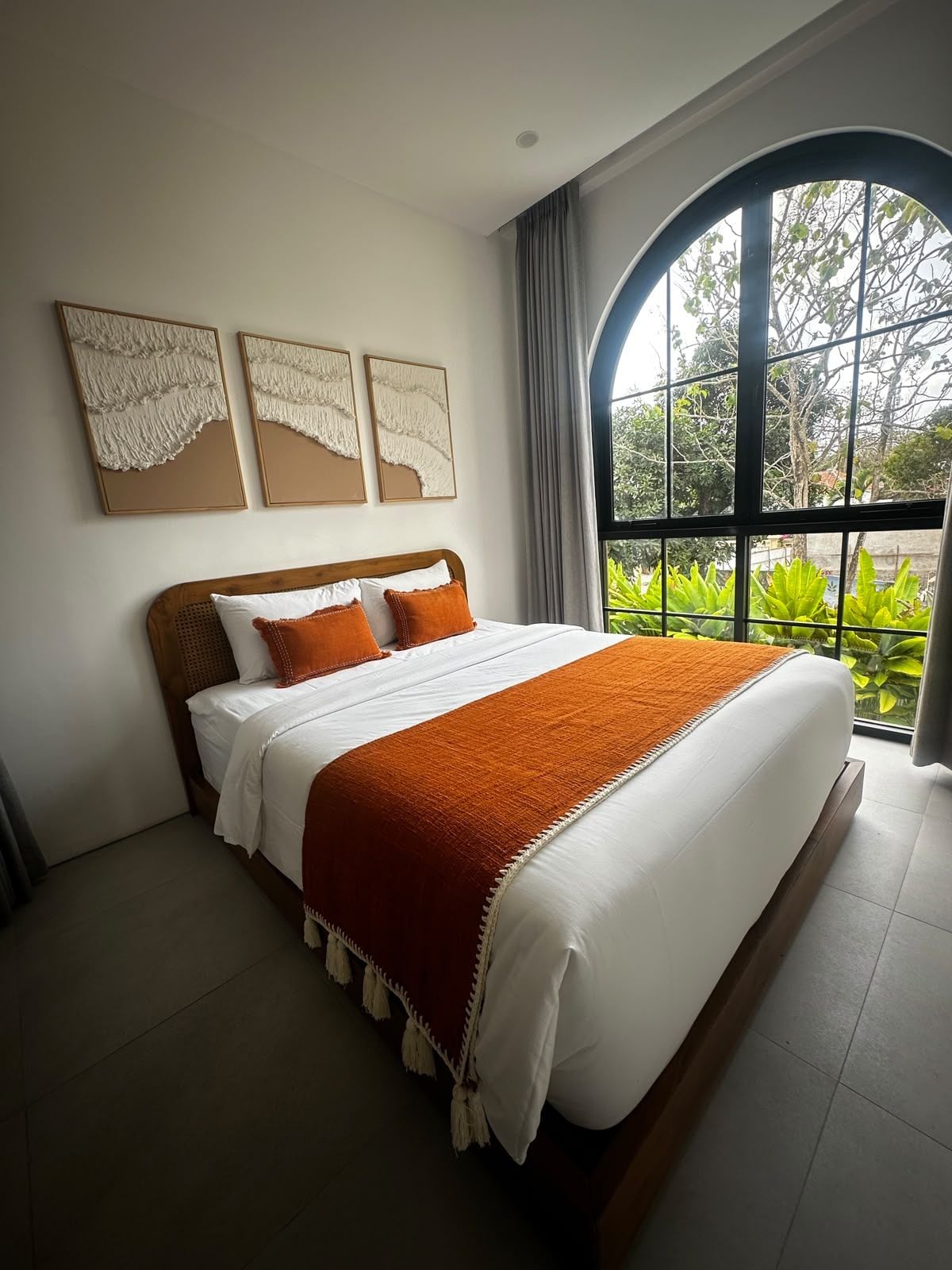 A bedroom with a large bed, a window with a view of greenery, and three wall-mounted artworks.