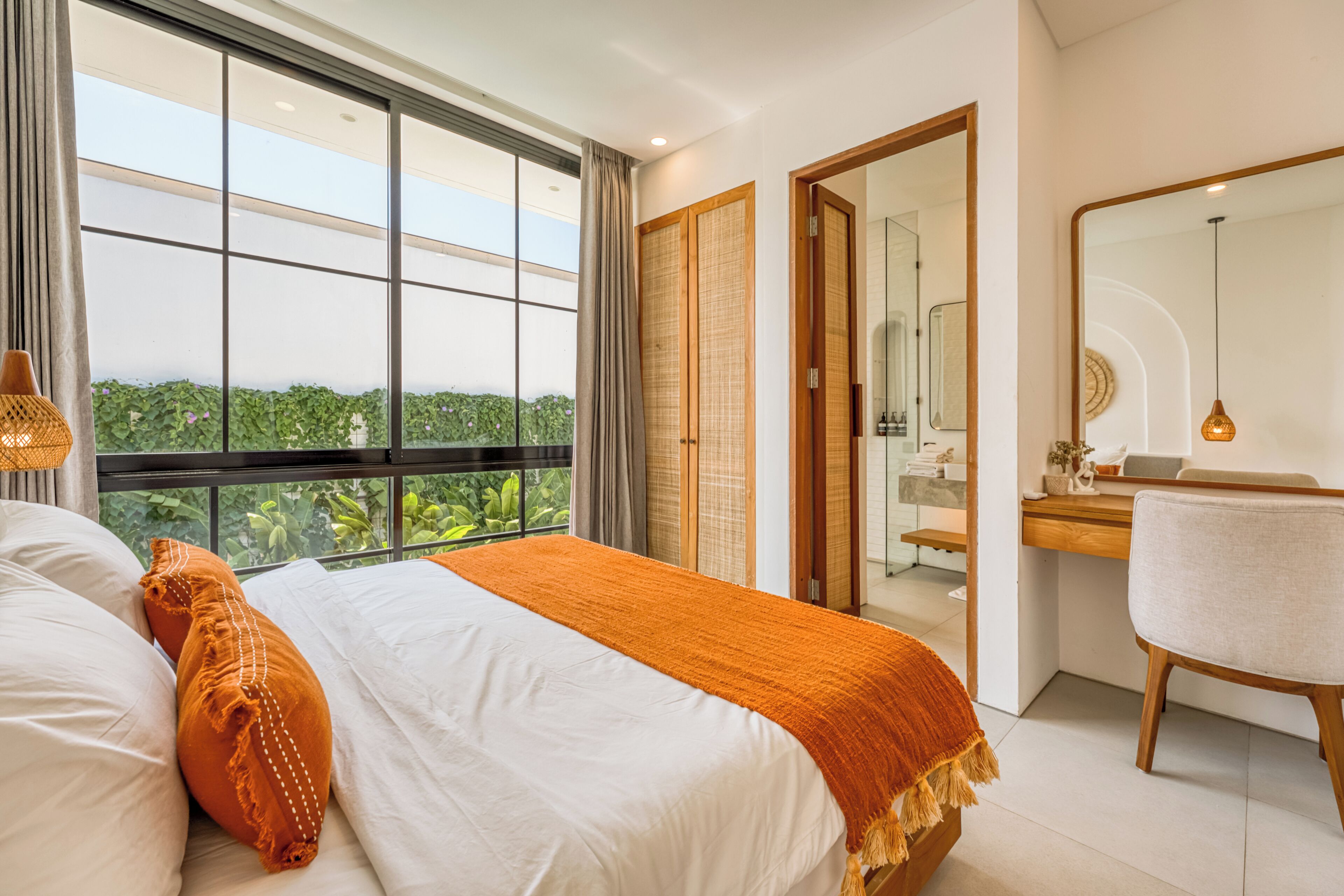 Una camera da letto con un'ampia finestra, un letto con biancheria da letto bianco e arancione, una scrivania in legno e uno specchio.