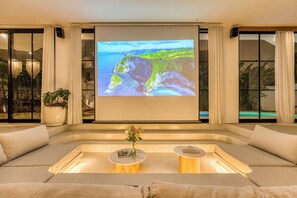 A modern living room with a large screen displaying a scenic landscape, comfortable seating, and a small table with a vase of flowers. - Senada Garden (Pecatu)