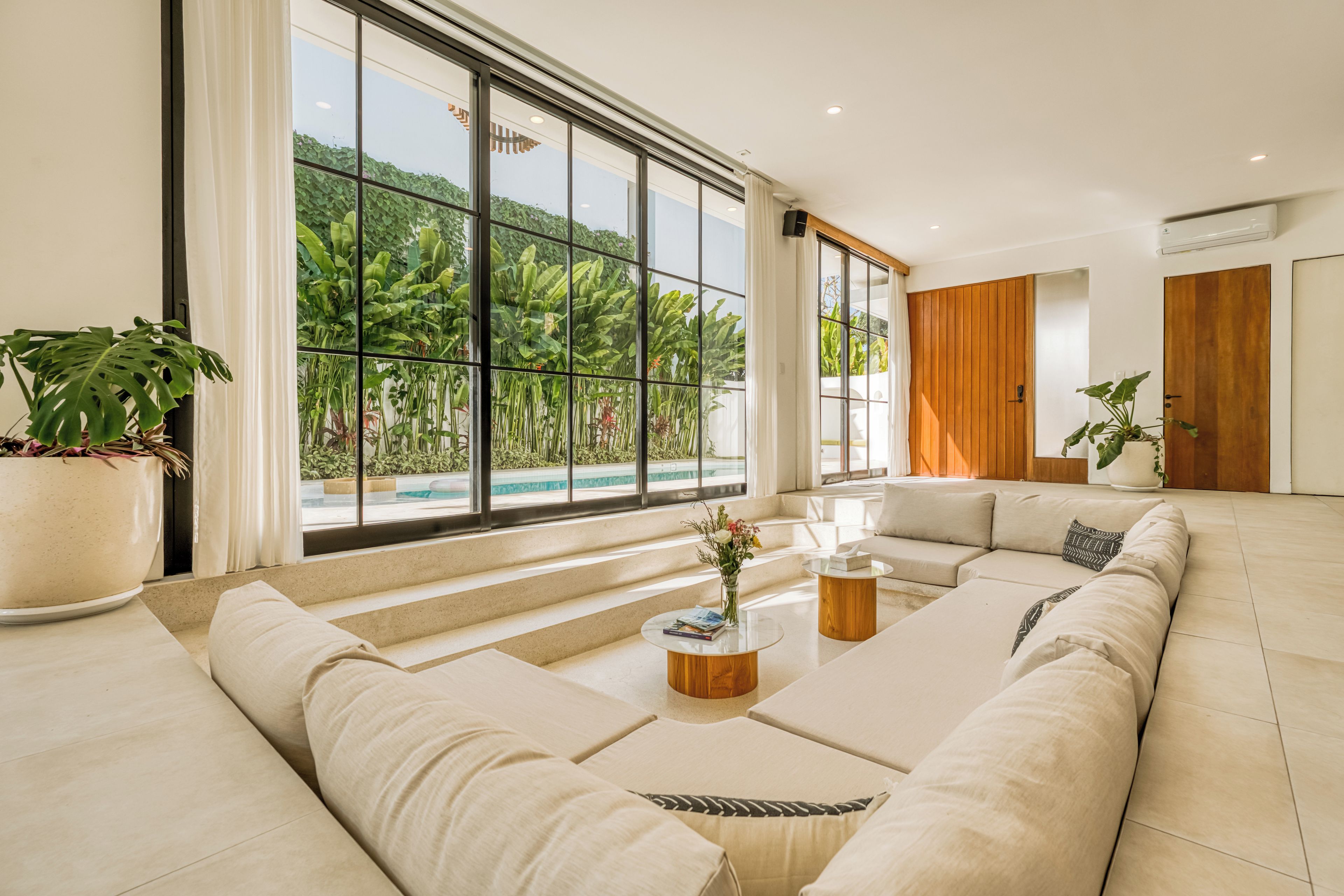 Un salon moderne doté d’une grande fenêtre, offrant une vue sur la piscine, et d’une porte en bois.