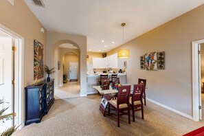 A dining area with a glass table and wooden chairs, a kitchen in the background, and artwork on the walls. - Snowbird-friendly resort condo with private balcony, pool & hot tub - near parks (Davenport)