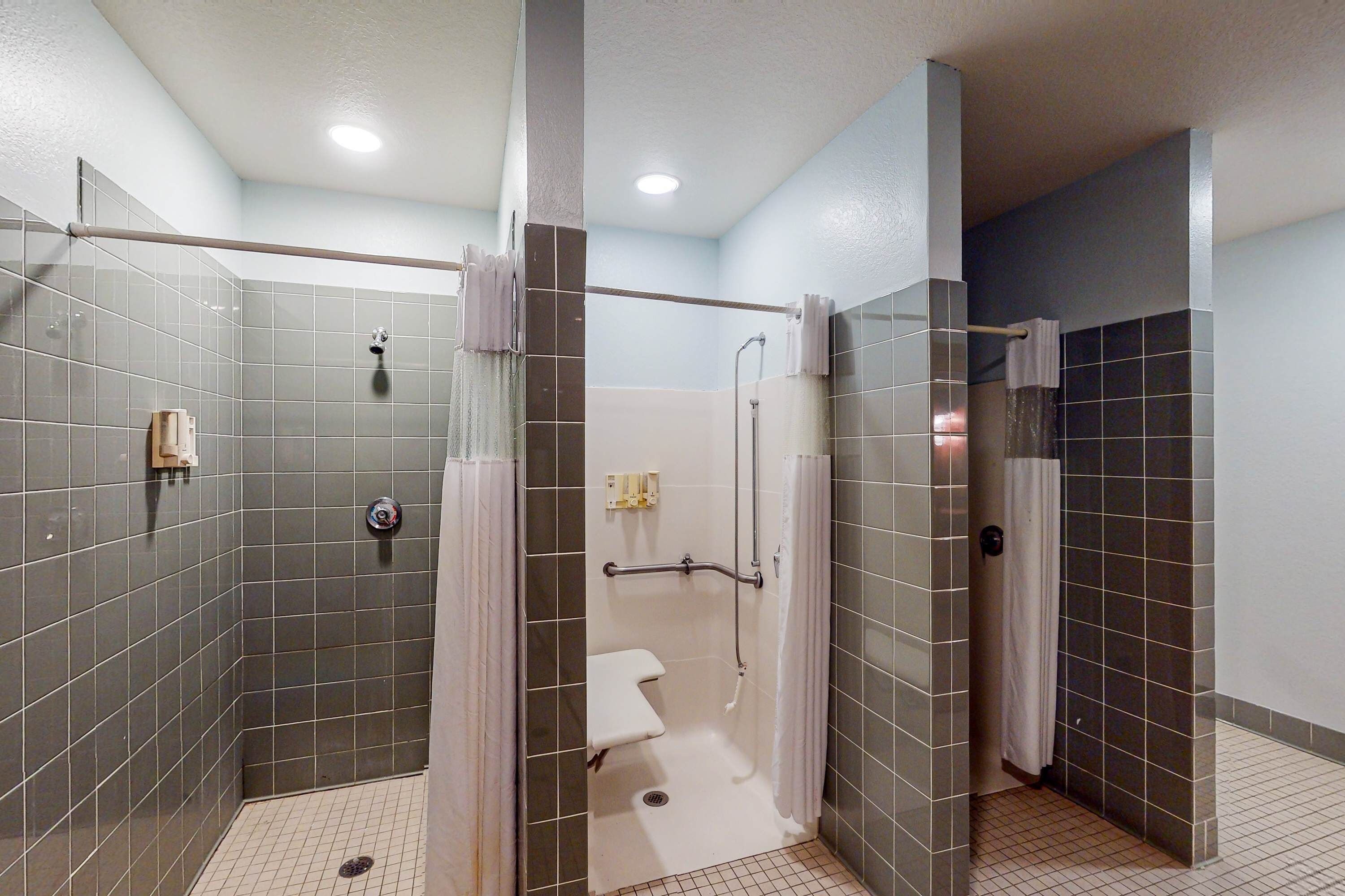 Une salle de bain moderne avec des murs recouverts de carreaux gris, des toilettes blanches, une douche avec une porte en verre et une barre d’appui.