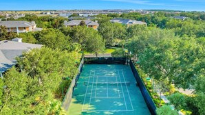 Ein Luftbild zeigt einen Tennisplatz, der von üppigem Grün und Wohnhäusern umgeben ist.