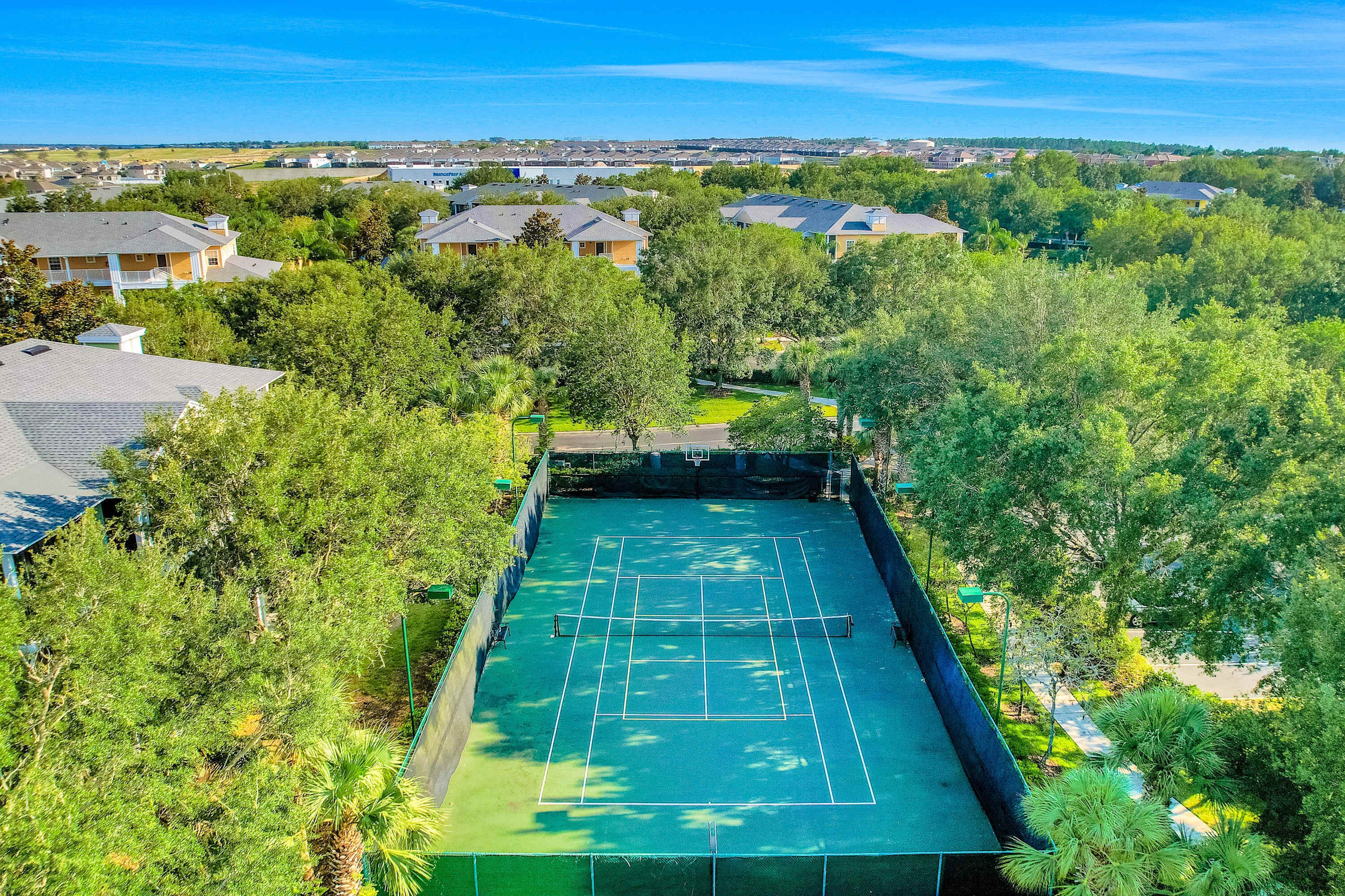 An aerial view of a tennis court surrounded by lush greenery and residential buildings.