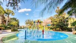 Un’area giochi d’acqua con una piscina circolare, circondata da palme e vegetazione.