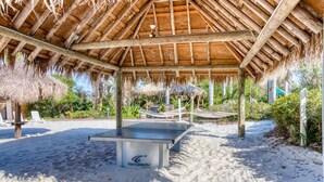 Ein Pavillon direkt am Strand mit einer Tischtennisplatte, umgeben von Sand und strohgedeckten Sonnenschirmen.