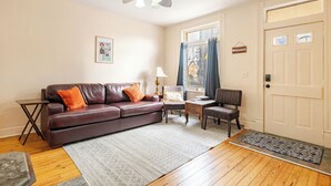 A living room with a brown leather sofa, a small table, a chair, a lamp, and a door. - Historic German Village Cottage - Perfect Downtown Location - Highly Walkable! (Columbus)