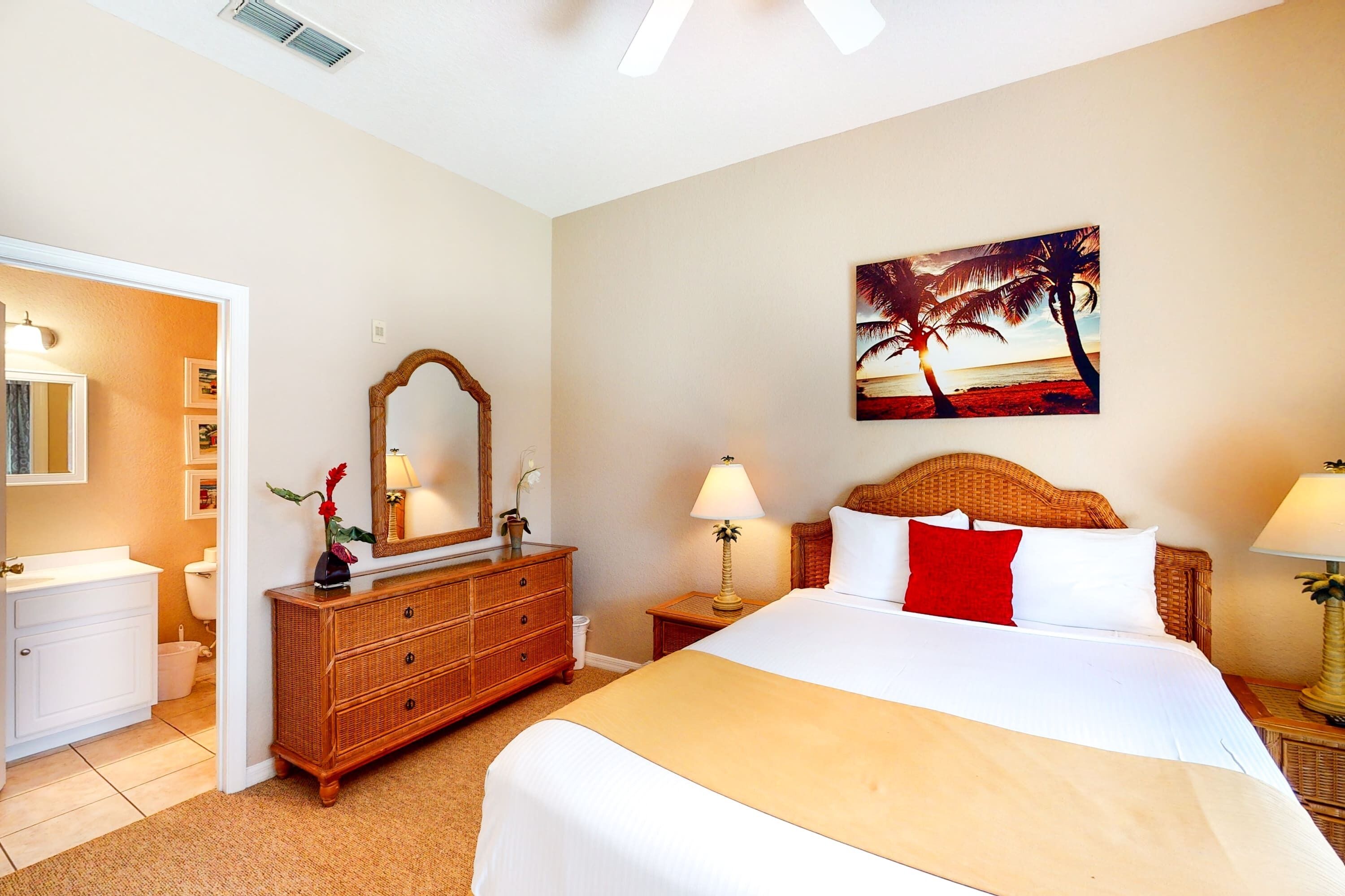 Habitación de hotel con cama, cómoda, espejo, un cuadro de una playa y un baño visible a través de una puerta abierta.
