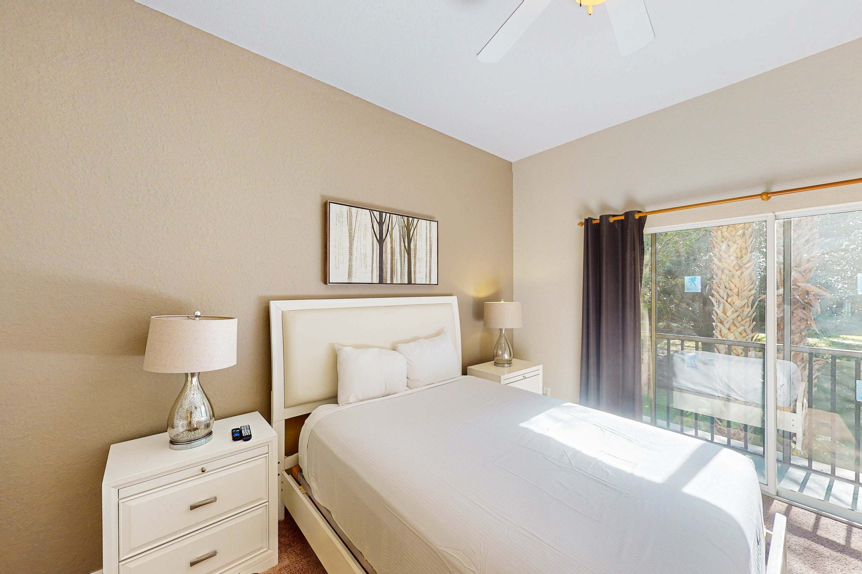 A hotel room with a large bed, a nightstand with a lamp, a small table with another lamp, a painting on the wall, and a balcony with a view of palm trees.