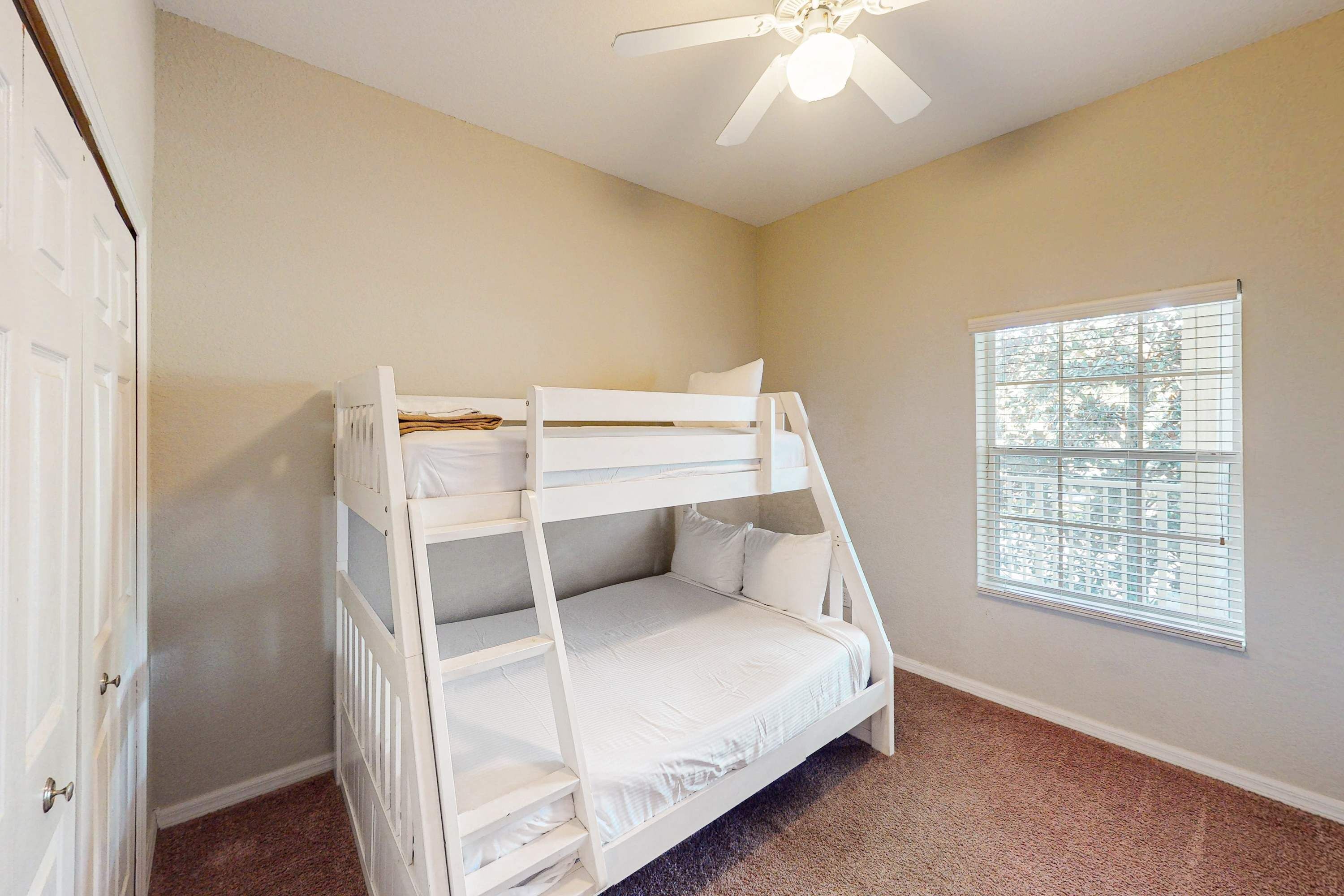 A bunk bed room with a ceiling fan, a window with blinds, and a door.