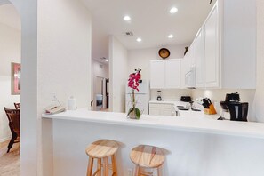 Eine moderne Küche mit weißen Schränken, einer Arbeitsplatte, zwei hölzernen Barhocker, einem Kühlschrank, einer Kaffeemaschine und einer Topfpflanze mit Orchideen.