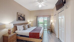Quarto com cama, abajures, ventilador de teto, televisão e janela com cortinas.