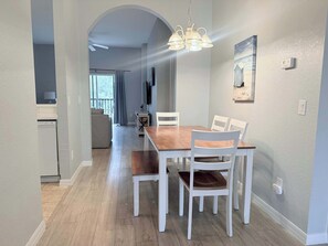 Un coin repas avec une table en bois et des chaises blanches, un ventilateur de plafond et un luminaire fixé au mur.