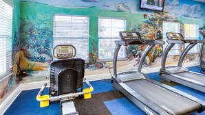 Ett gym med löpband, en motionscykel och en roddmaskin. Det finns fönster med persienner, en väggmålning som föreställer en undervattensscen och en tv som är monterad på väggen.