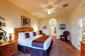 Une chambre d’hôtel avec un grand lit, une tête de lit en bois, deux tables de chevet avec des lampes, un ventilateur de plafond et une vue sur le couloir.