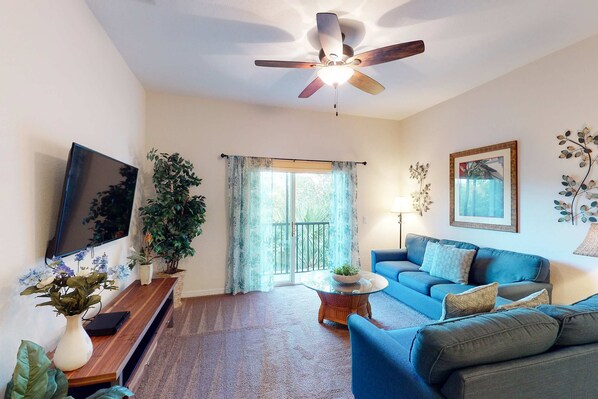 Un salon avec un ventilateur de plafond, un téléviseur, un canapé, une table basse et une plante.