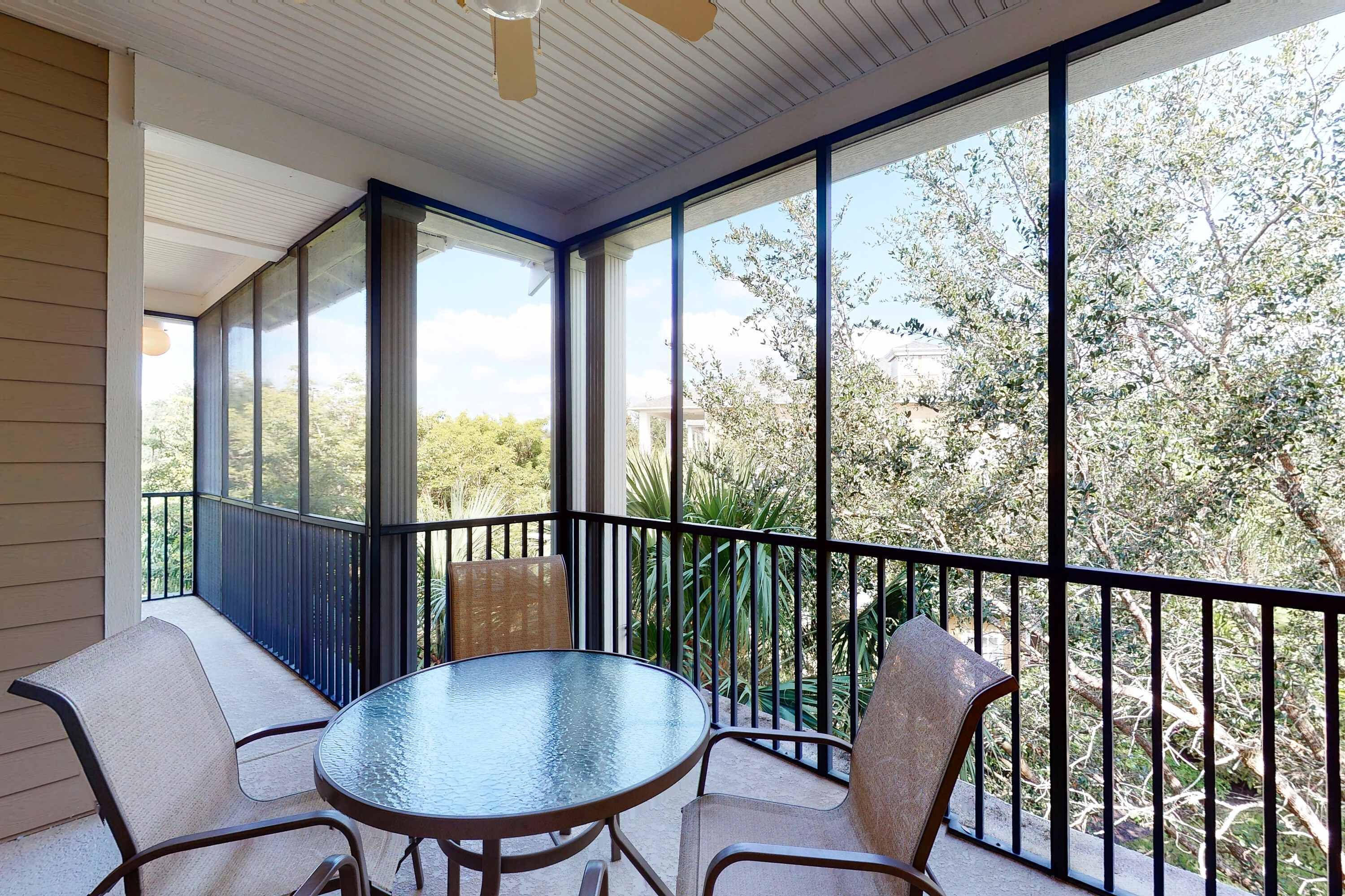 A covered balcony with a glass table and chairs, offering a view of trees and a building in the background.