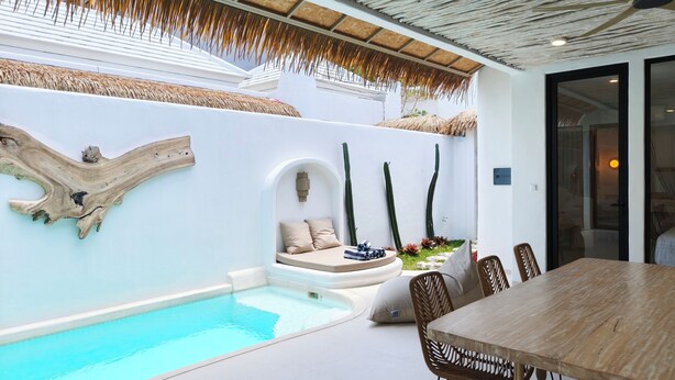 Un espace piscine extérieur moderne, avec une table et des chaises en bois, un coin salon avec des coussins et un mur décoratif orné de cactus.
