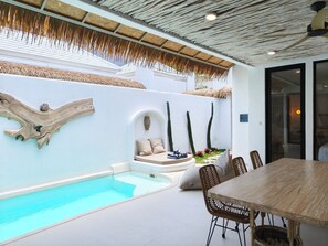 Un espace piscine extérieur moderne, avec une table et des chaises en bois, un coin salon avec des coussins et un mur décoratif orné de cactus.