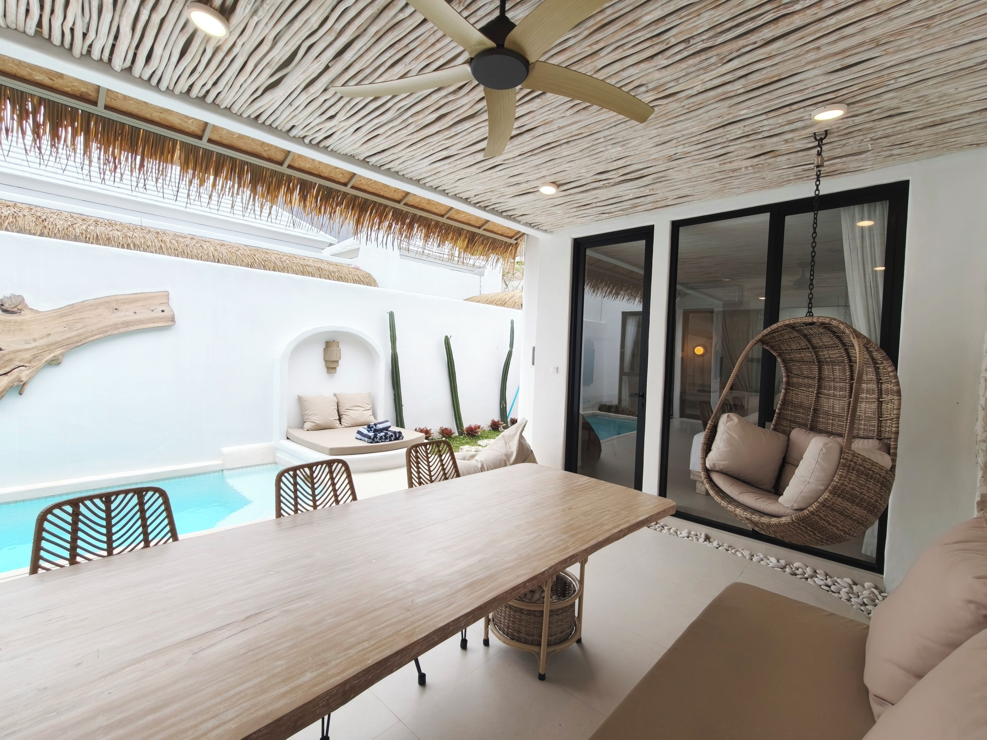 A modern room with a wooden dining table, a hanging chair, a small seating area with cushions, and a view of a pool through a glass door.