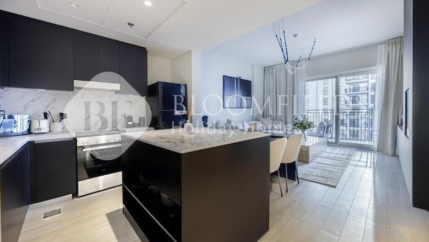A modern kitchen with dark cabinetry, a central island, and a dining area with a view to the outside. - Bloomfields Formula 1br In Water (Abu Dhabi)