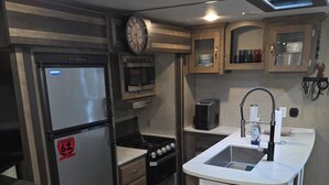 Uma cozinha compacta com geladeira, micro-ondas, fogão, pia e um relógio na parede.