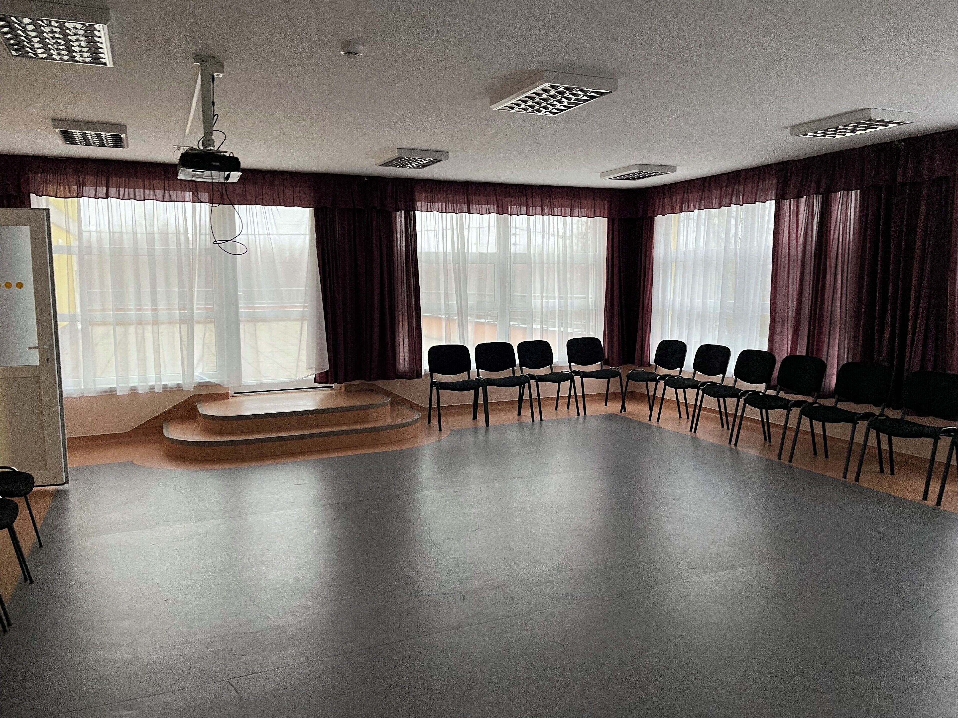 Een vergaderzaal met een podium, rijen stoelen en grote ramen voorzien van gordijnen.
