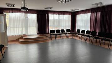 Een vergaderzaal met een podium, rijen stoelen en grote ramen voorzien van gordijnen.
