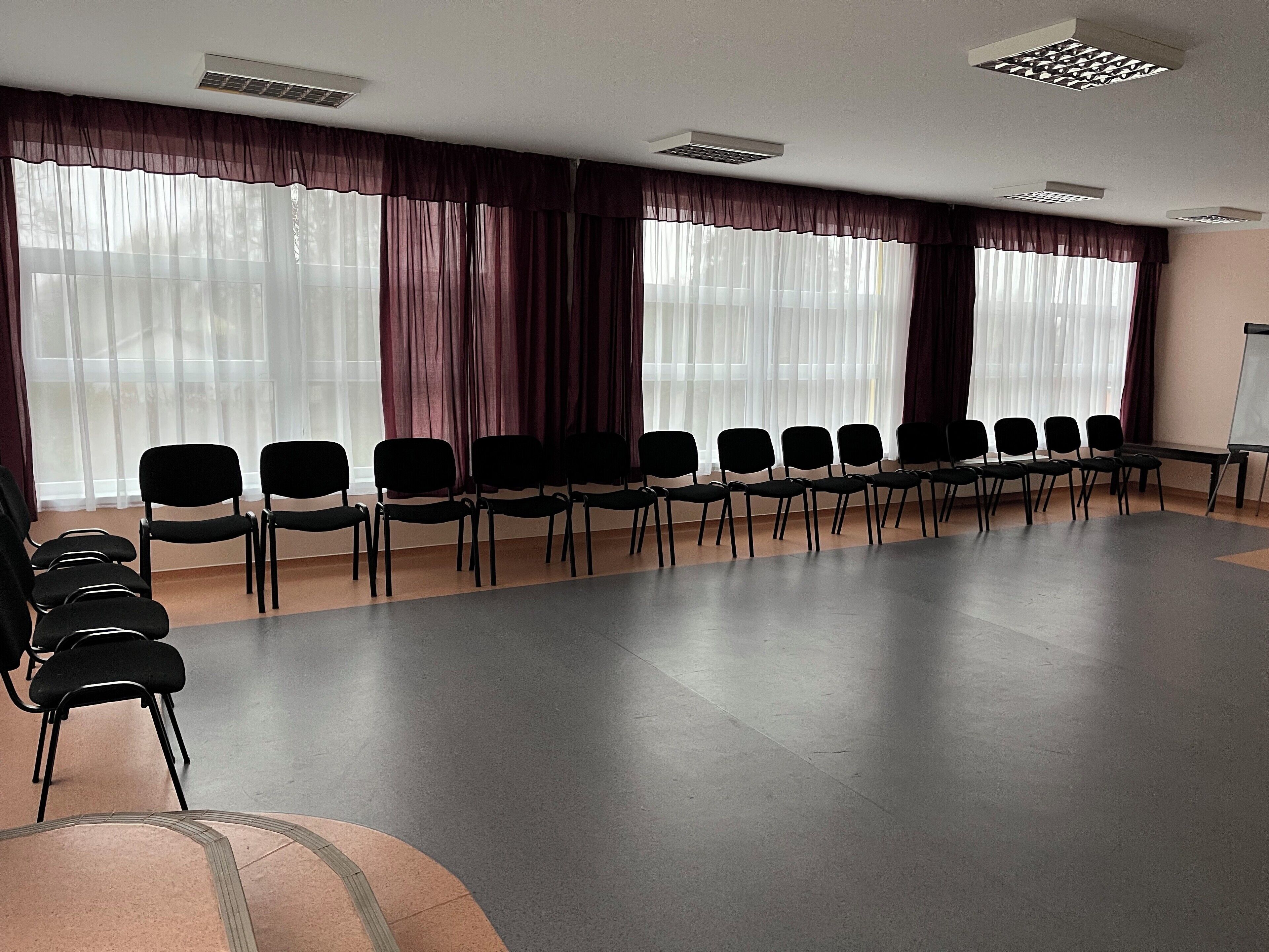 Un salón de conferencias con filas de sillas, ventanas amplias con cortinas translúcidas y una pizarra.