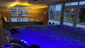 Un bain à remous avec un éclairage bleu, une vue sur une piscine à l’extérieur et un mur carrelé avec une fenêtre.