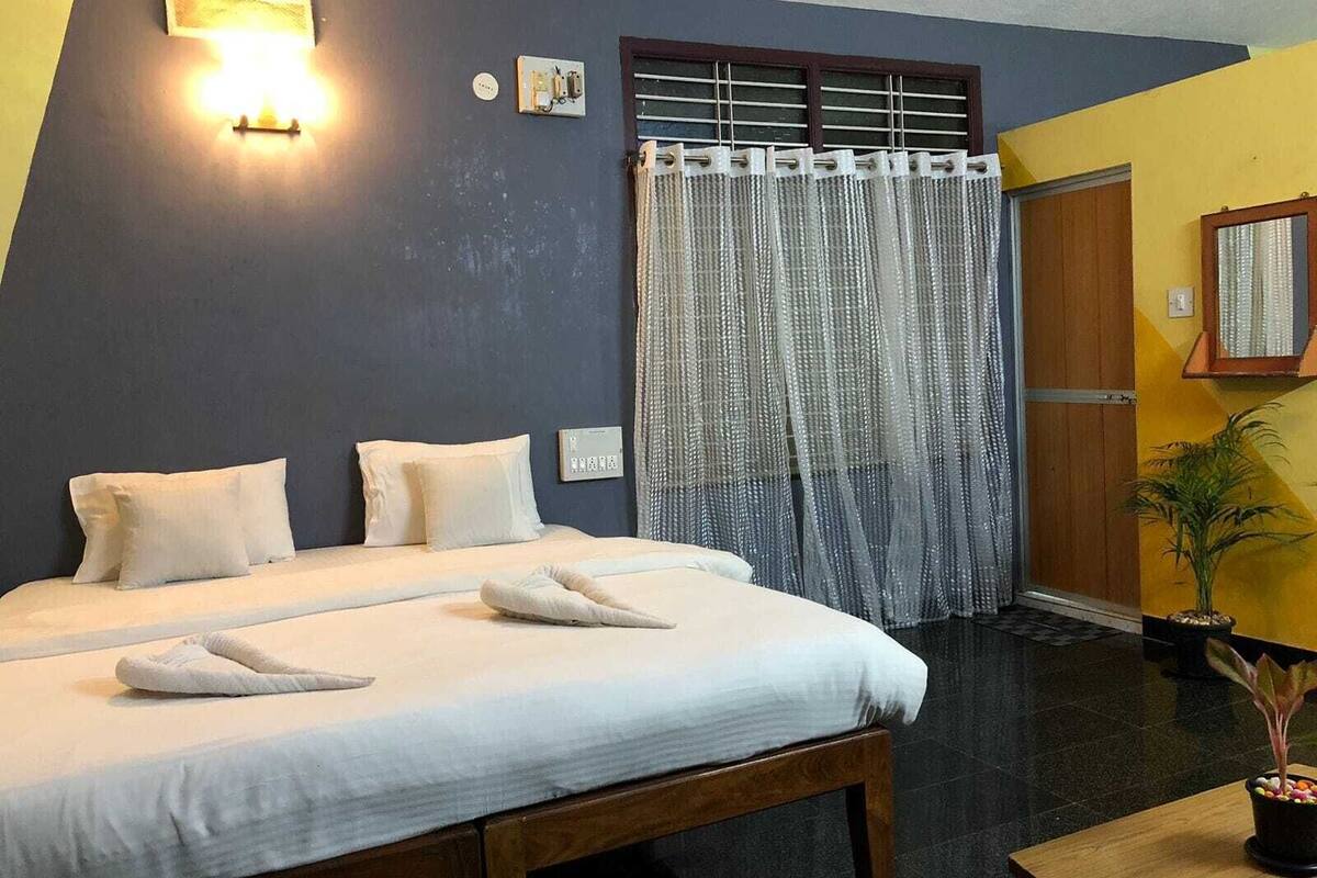 A hotel room with a bed, a bedside table, a potted plant, and a window with curtains.