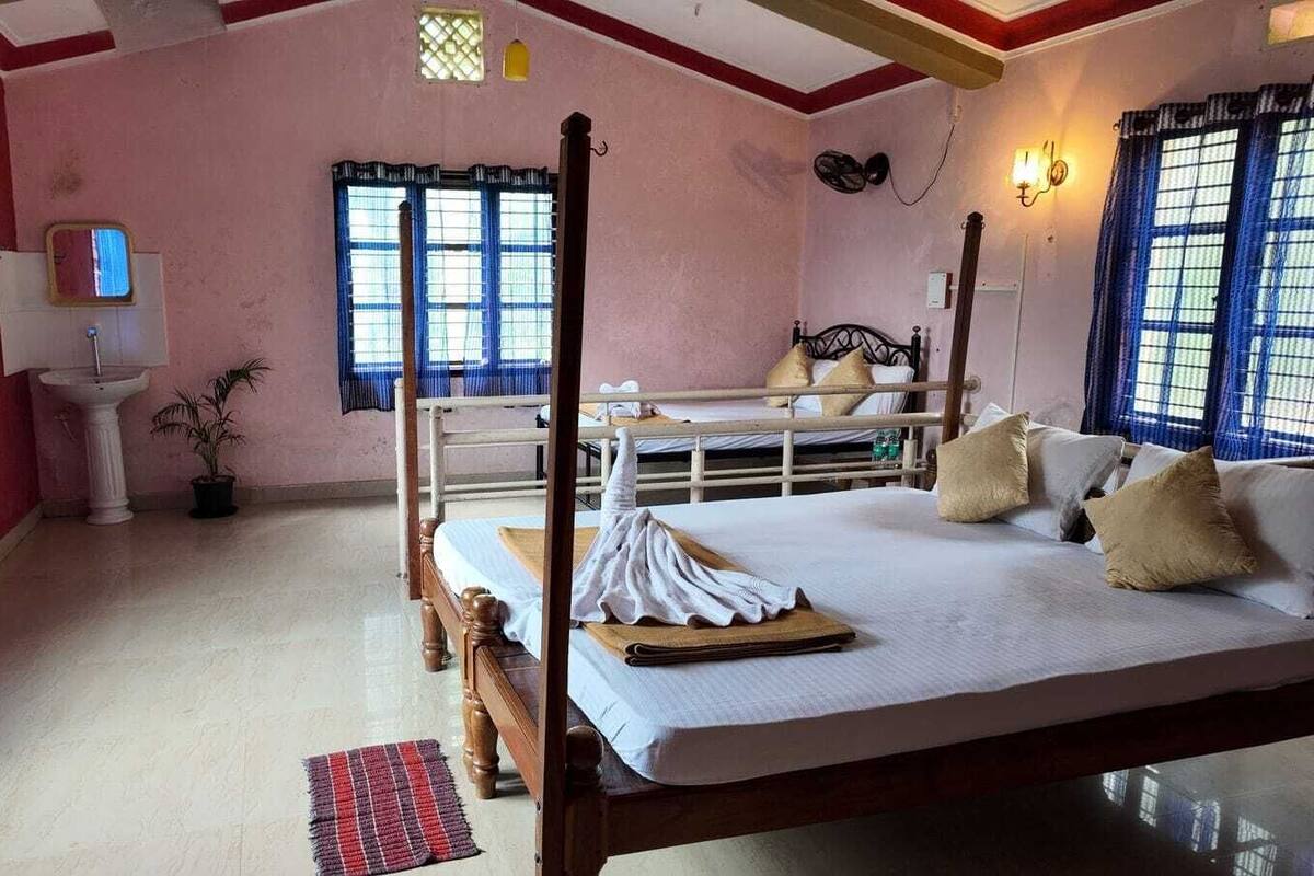 A four-poster bed with a white mattress and beige pillows, a small bedside table with a lamp, a potted plant, and a window with blue curtains.