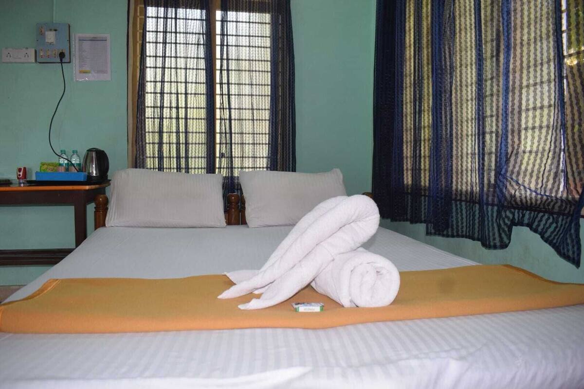 A hotel room with a bed, pillows, a rolled towel, a bedside table with a kettle, and a window with curtains.
