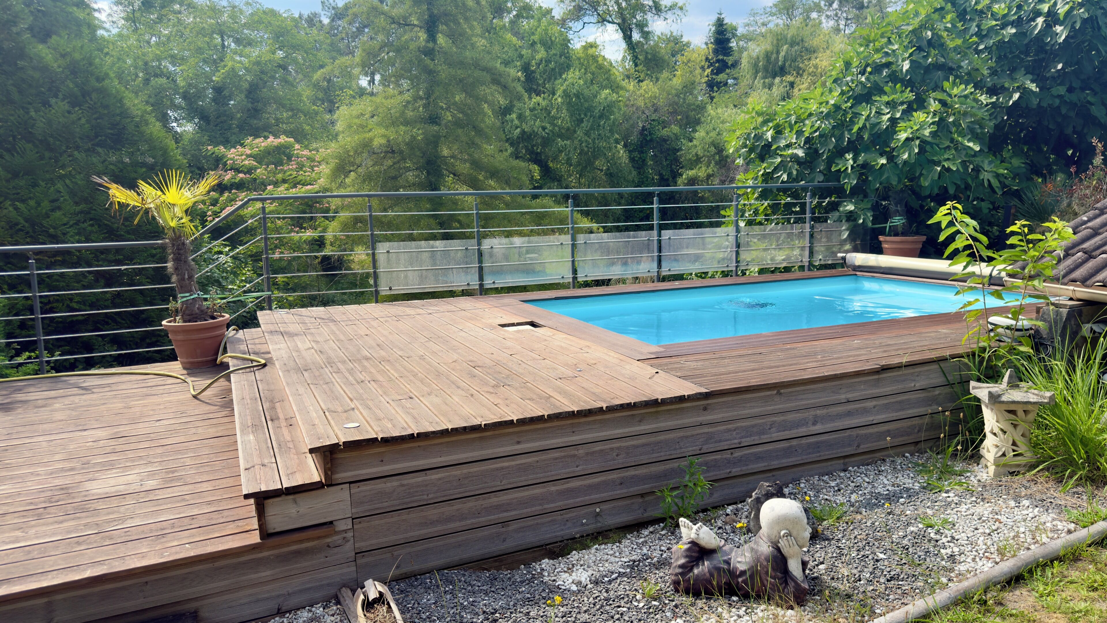 Une terrasse en bois avec une piscine, entourée d’une clôture en métal et de verdure.