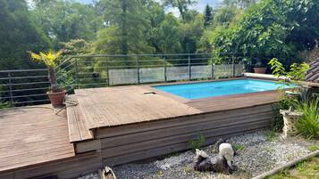 Une terrasse en bois avec une piscine, entourée d’une clôture en métal et de verdure.