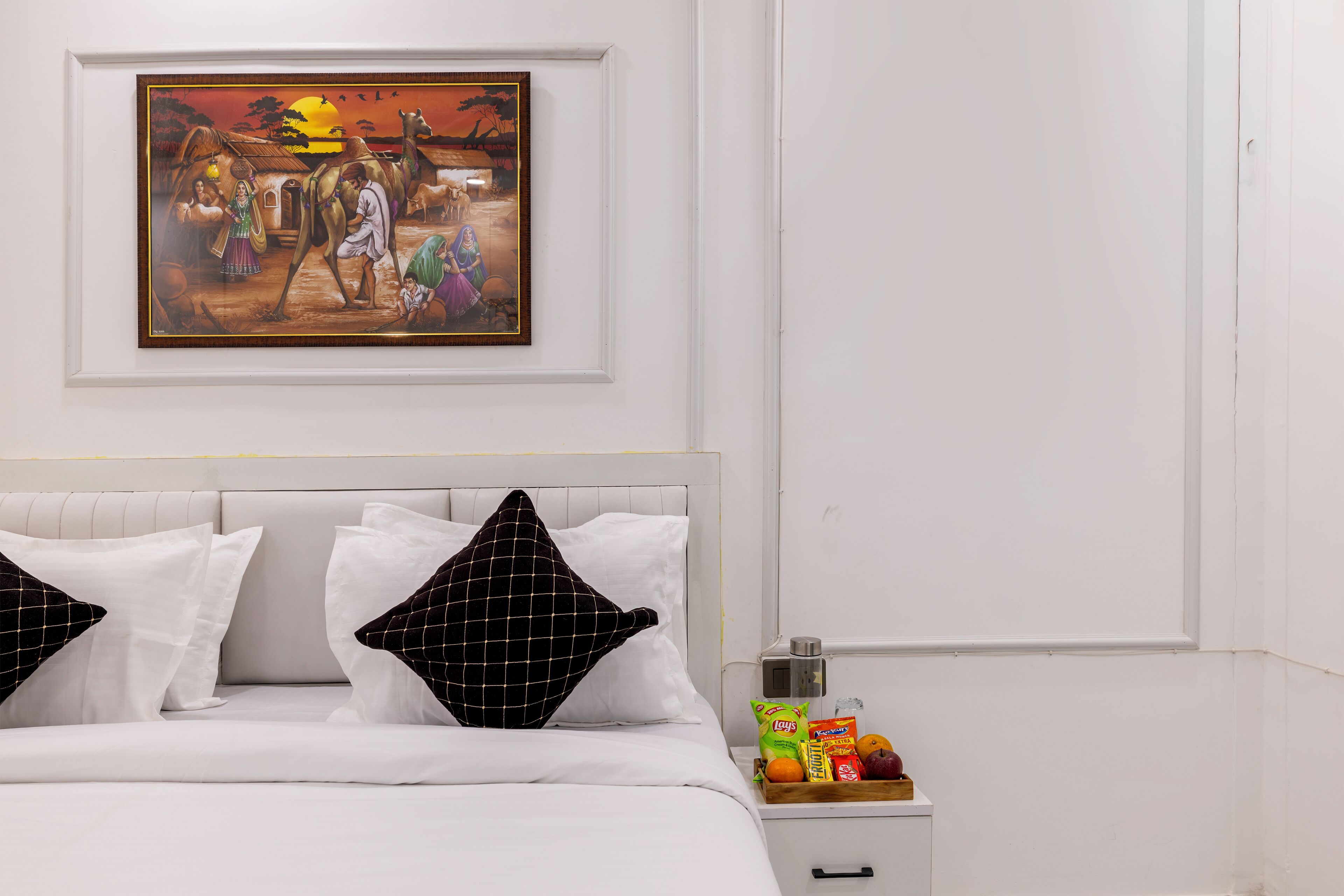 Una habitación de hotel con una cama, almohadas, una mesita de noche con aperitivos y un cuadro en la pared.