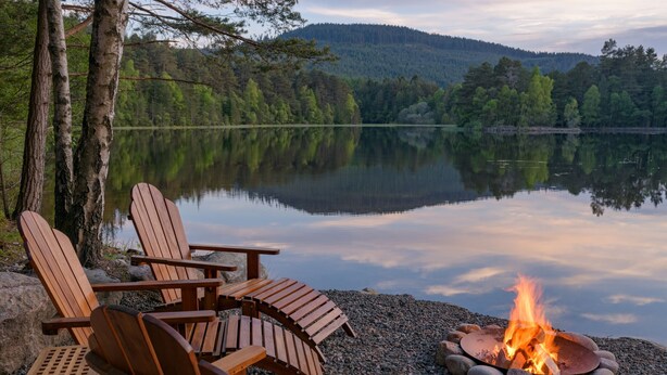 A lakeside setting with wooden Adirondack chairs and a campfire. - The Queen's Hut - Idyllic Log Cabin, originally built for Queen Mary (Aboyne)